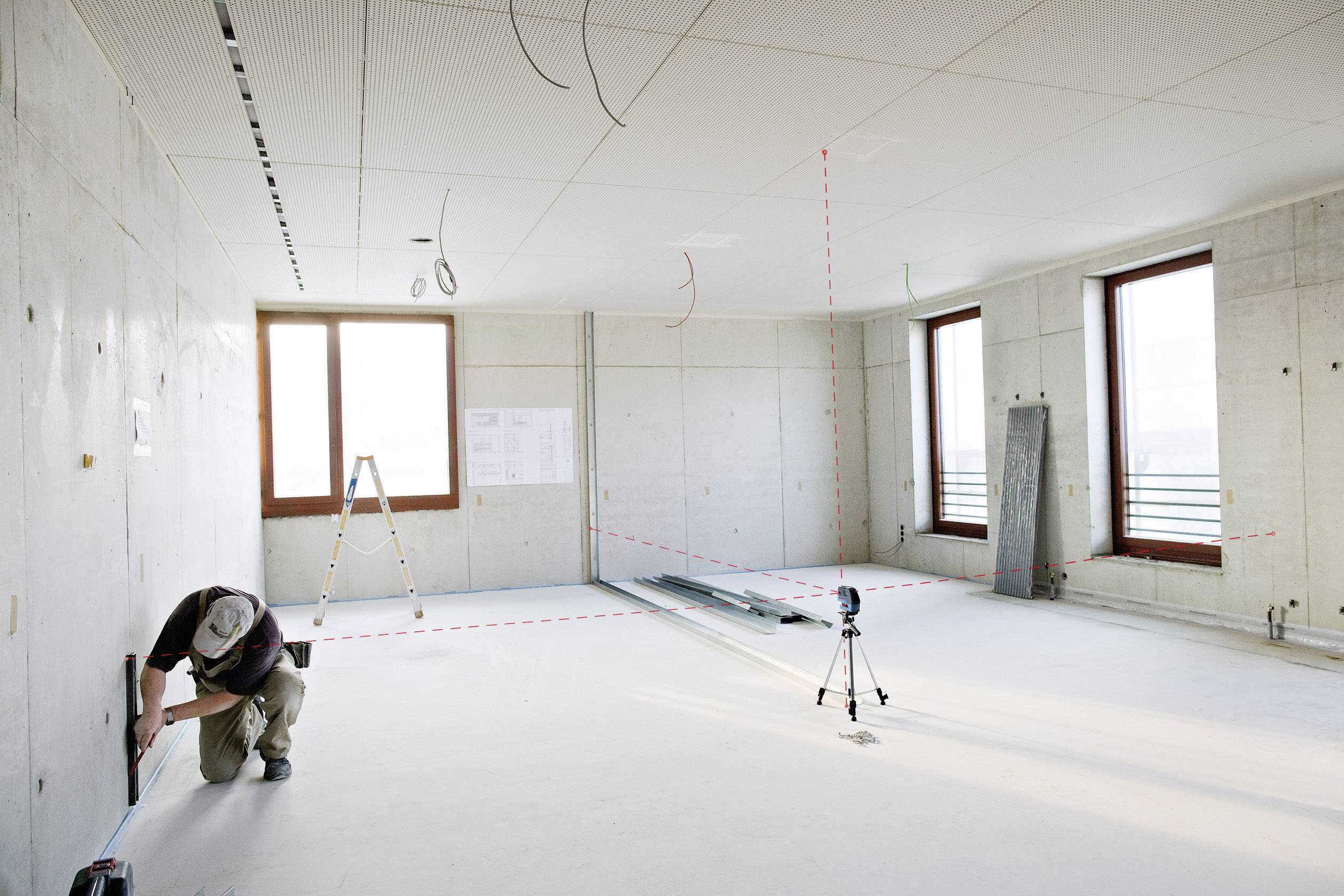 Ein Handwerker misst in einem leeren, unfertigen Raum mit Laserwerkzeugen an der Wand. Gerüste und Baumaterialien sind sichtbar.