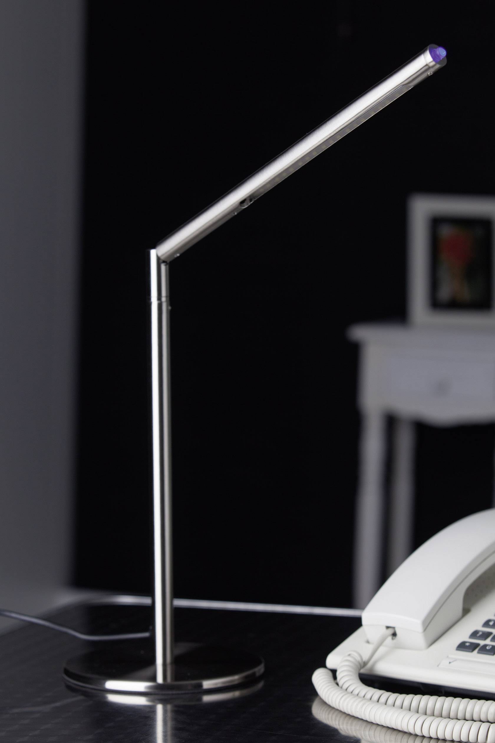 Metallische Schreibtischlampe mit schlankem Design, beleuchtet einen modernen Arbeitsplatz; im Hintergrund ein Telefon auf einem Tisch.