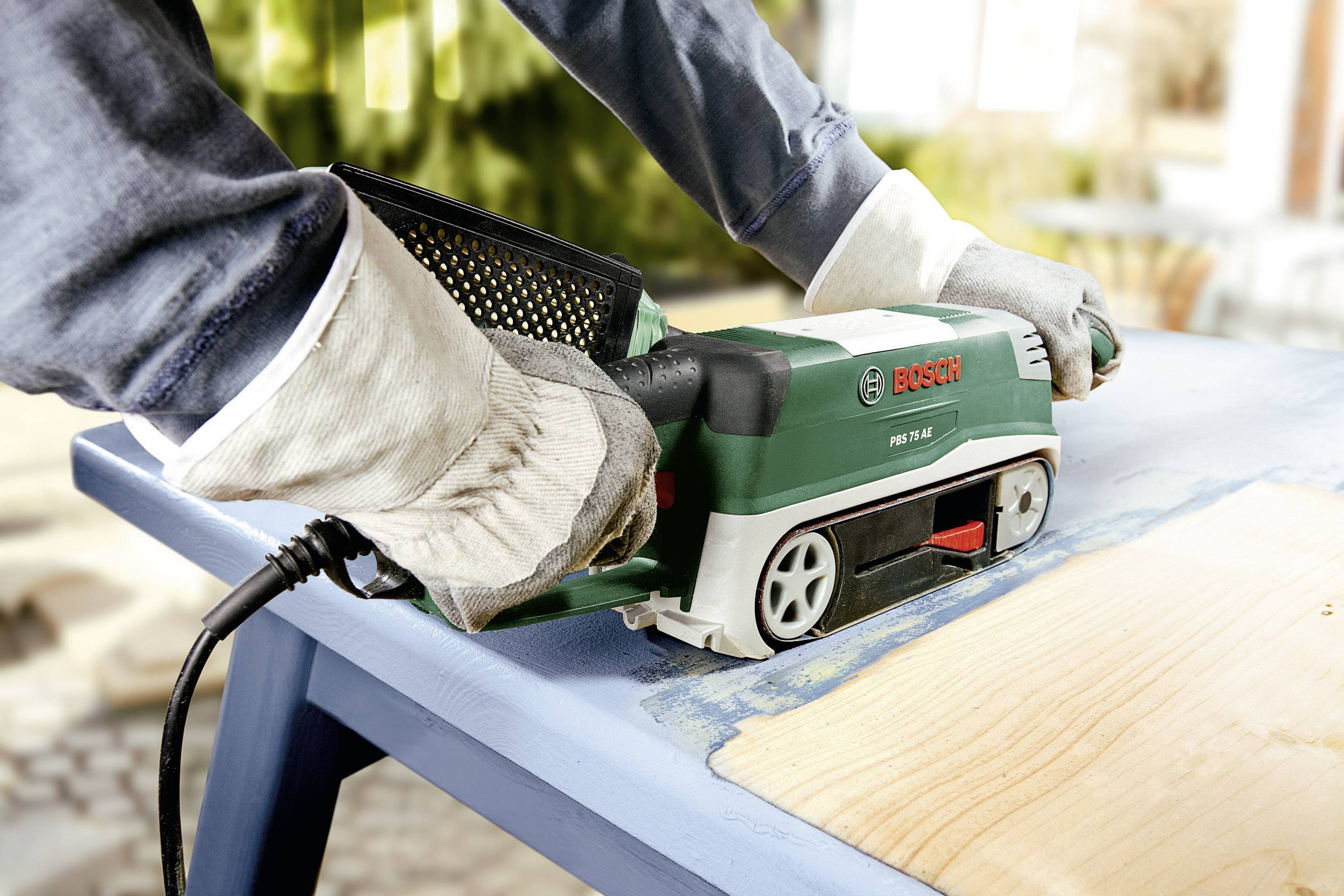 'Eine Person schleift mit einer Bosch-Bandschleifmaschine Holz auf einem Tisch. Sie trägt Arbeitshandschuhe und bearbeitet eine helle Holzplatte.'