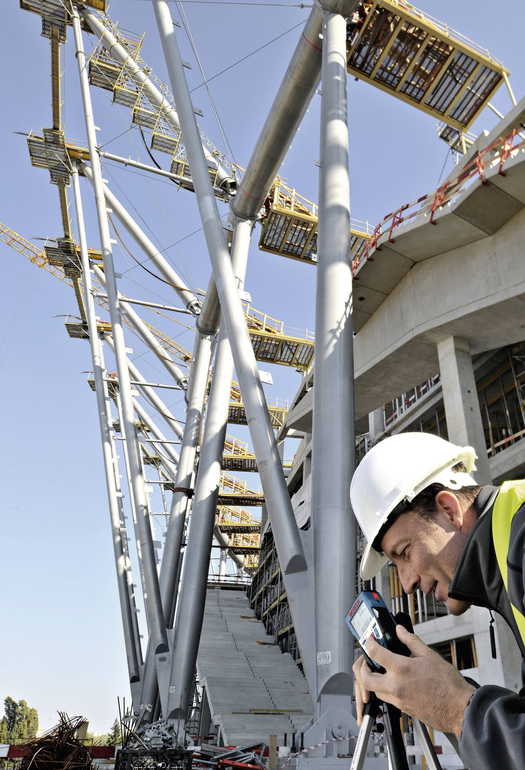 Ein Bauarbeiter mit Helm misst mithilfe eines Geräts an einer modernen Stahlkonstruktion auf einer Baustelle.