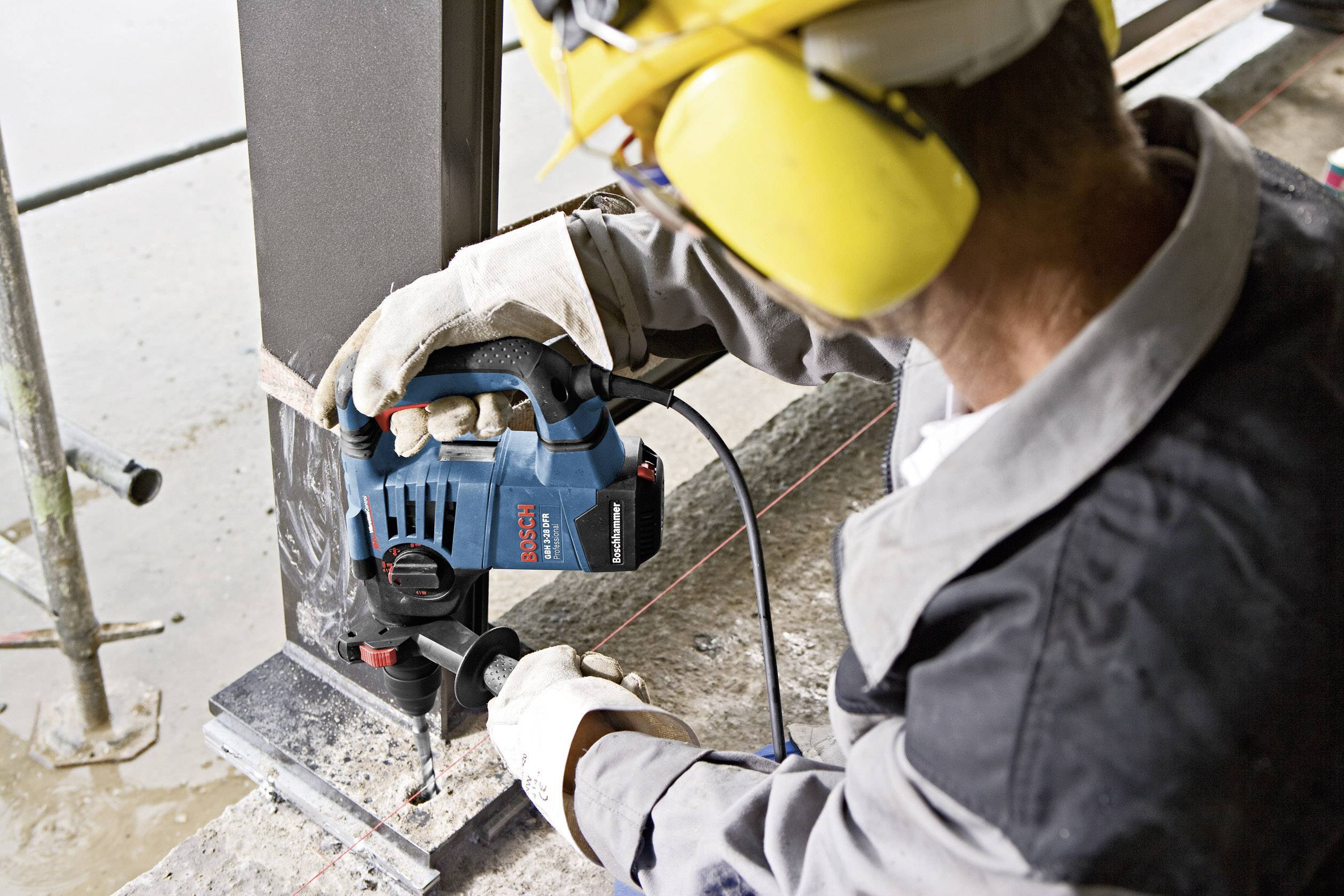 Eine Person in Schutzkleidung arbeitet mit einer blauen Stichsäge an einem Metallpfosten auf einer Baustelle. Schutzbrille und Gelb-Schallschutz sind sichtbar.