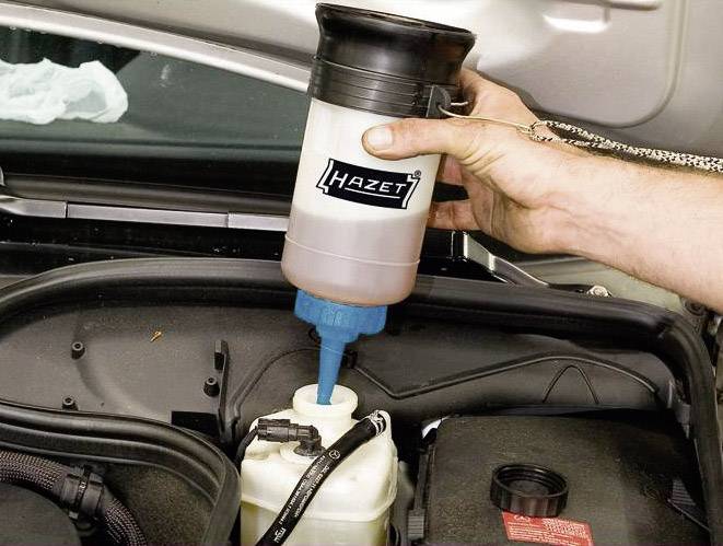 Eine Hand füllt Bremsflüssigkeit mit einer Hazet-Flasche in den Behälter eines Autos. Der Motorraum ist sichtbar.