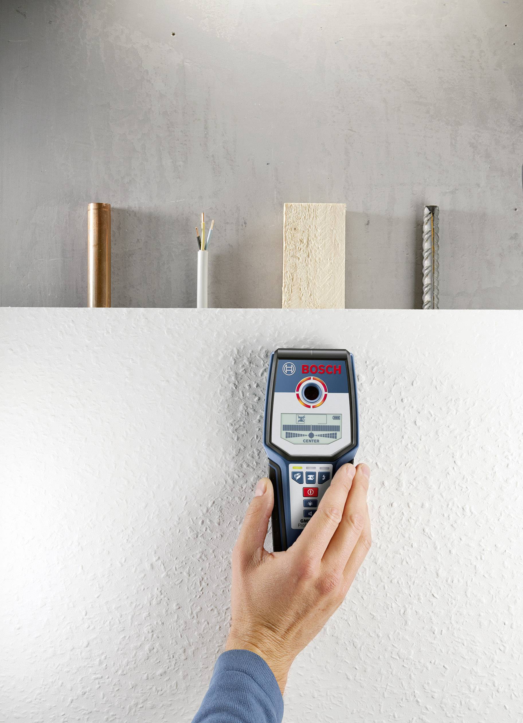 Eine Hand hält ein elektronisches Messgerät an einer weißen Wand. Auf der Wand sind dahinter Rohre unterschiedlicher Materialien sichtbar.