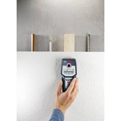 Eine Hand hält ein elektronisches Messgerät an einer weißen Wand. Auf der Wand sind dahinter Rohre unterschiedlicher Materialien sichtbar.