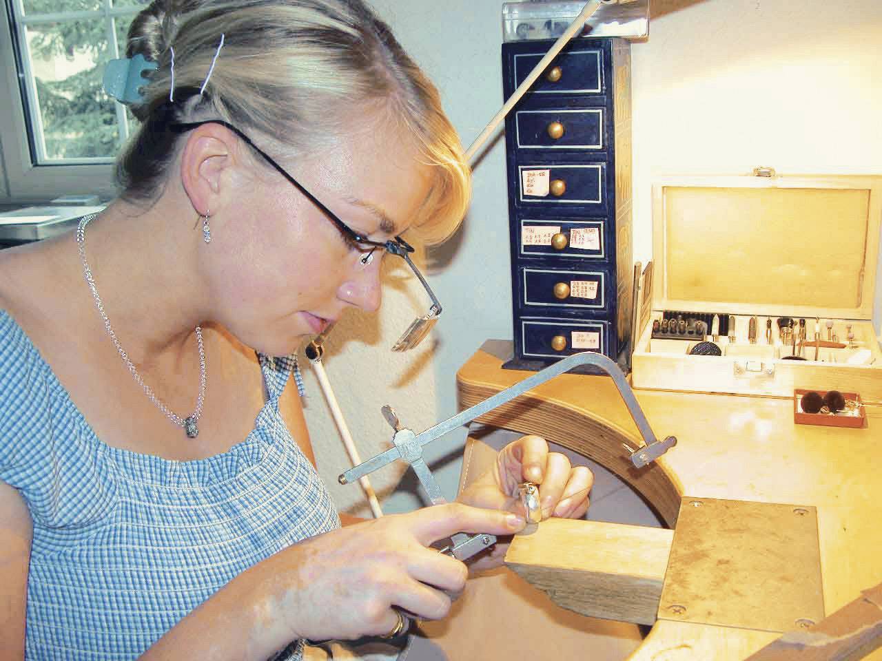 Eine Frau mit Brille arbeitet konzentriert an einem Schmuckstück in einer Werkstatt. Sie hält Werkzeuge in der Hand.