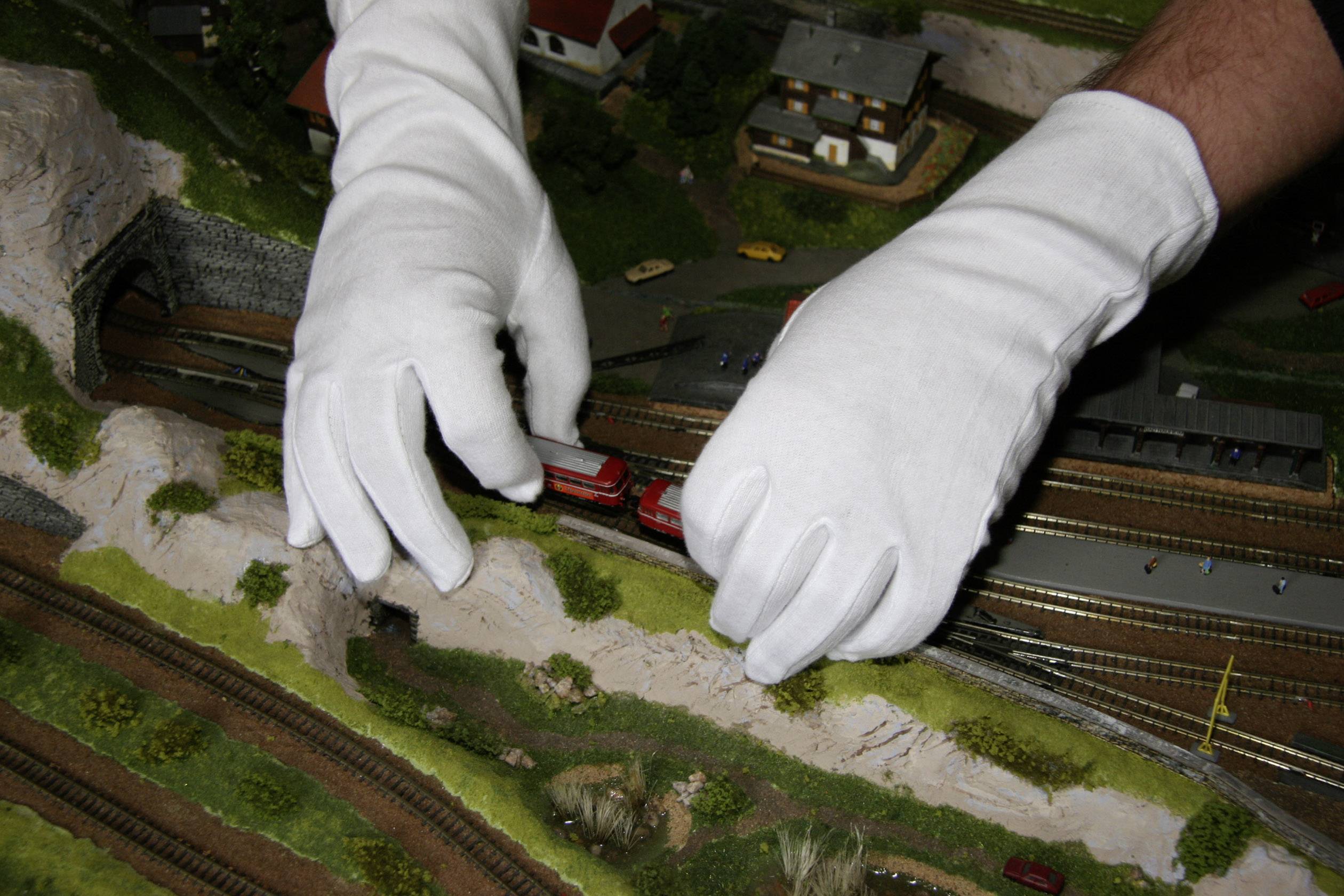 Zwei Hände in weißen Handschuhen greifen nach einer Spielzeugeisenbahn auf einer Modelleisenbahnlandschaft.