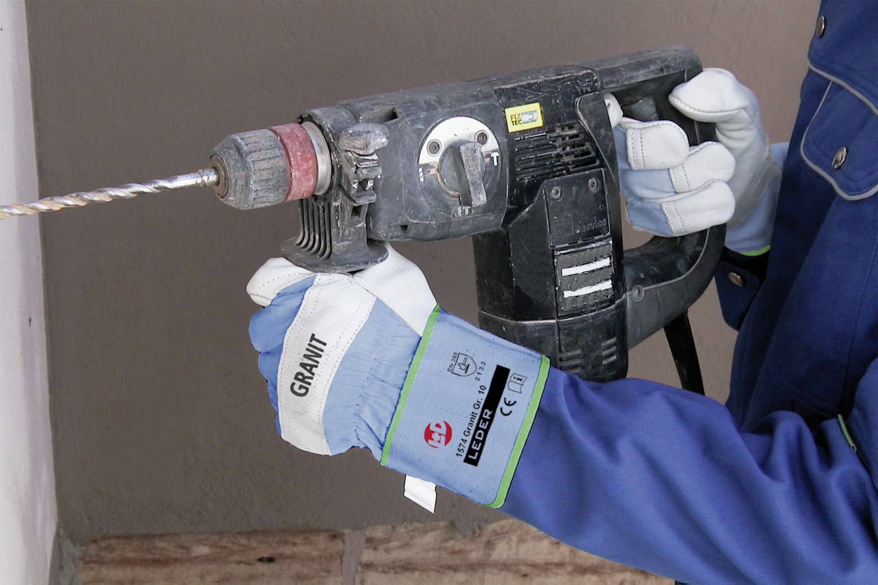 Eine Person in blauer Arbeitskleidung hält eine elektrische Bohrmaschine und bohrt in eine Wand. Handschuhe schützen die Hände.