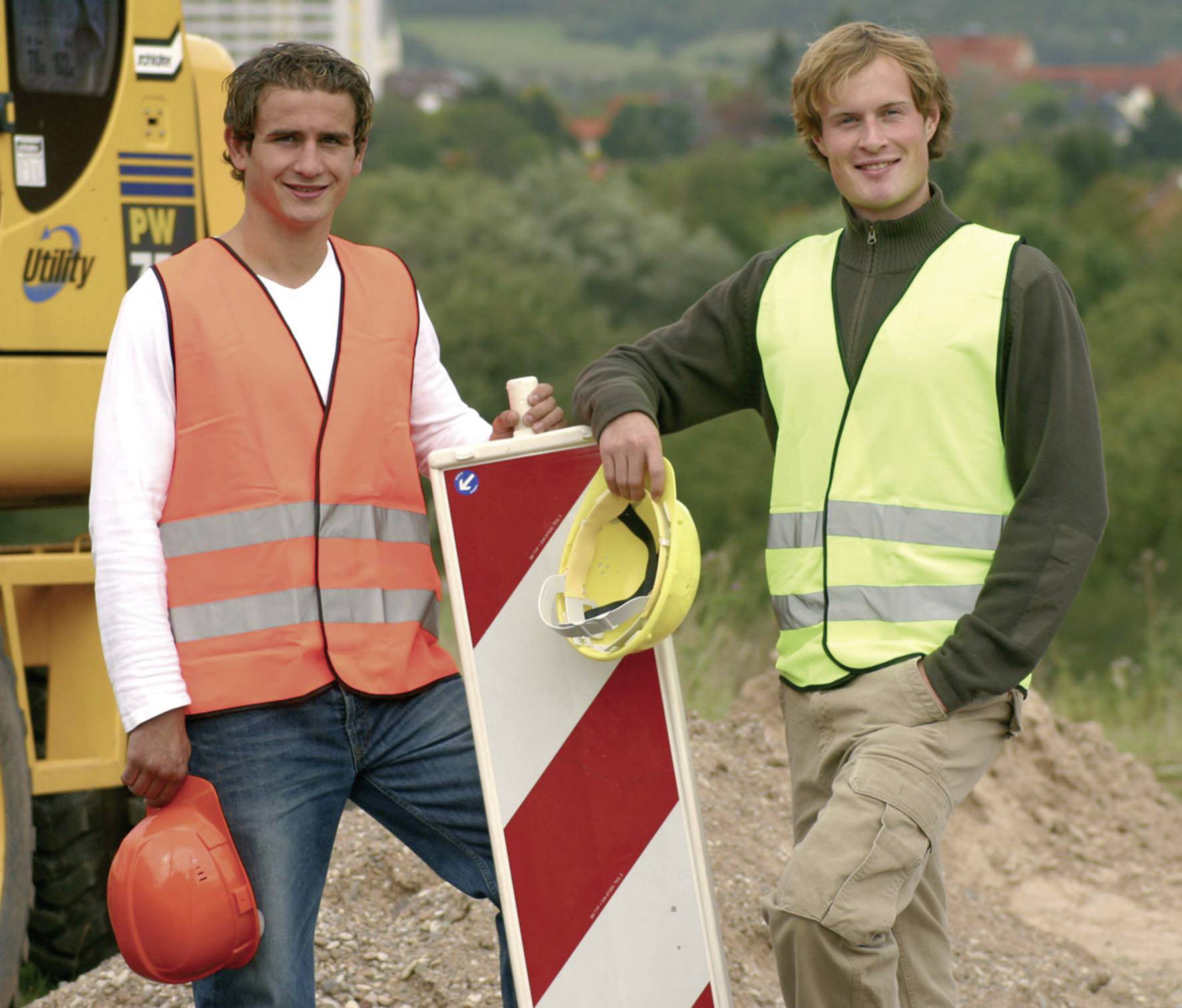 Zwei Personen in Schutzwesten stehen an einer Baustelle, halten Bauhelme und lehnen an einem Verkehrsschild. Hintergrund zeigt Landschaft.