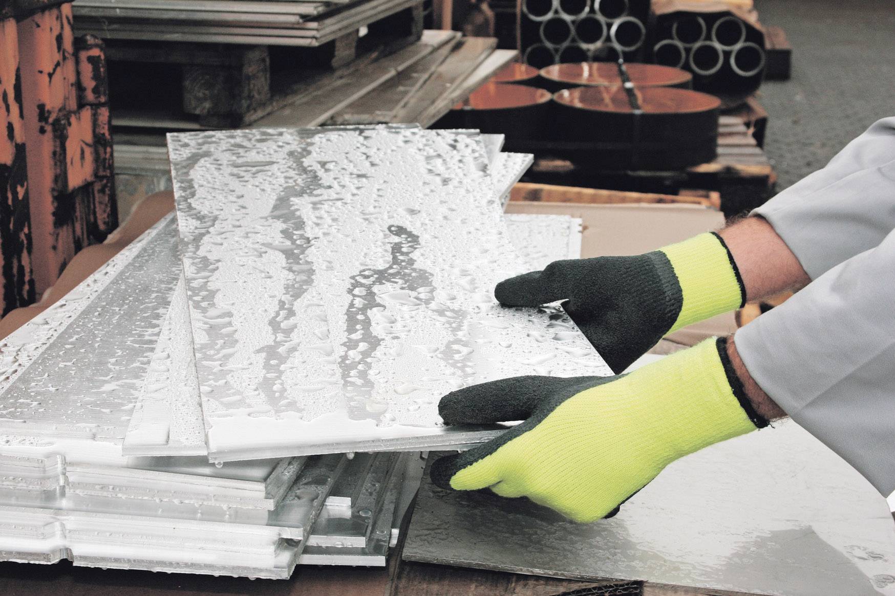 Eine Person mit gelben Handschuhen hebt metallene Bleche in einer Werkstatt oder Fabrik. Stapel von Metallplatten im Hintergrund.