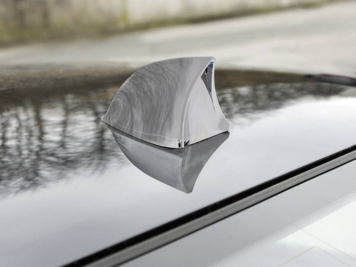 Eine silberne Sharkfin-Antenne ist auf dem Dach eines Autos montiert. Sie reflektiert leicht die Umgebung.