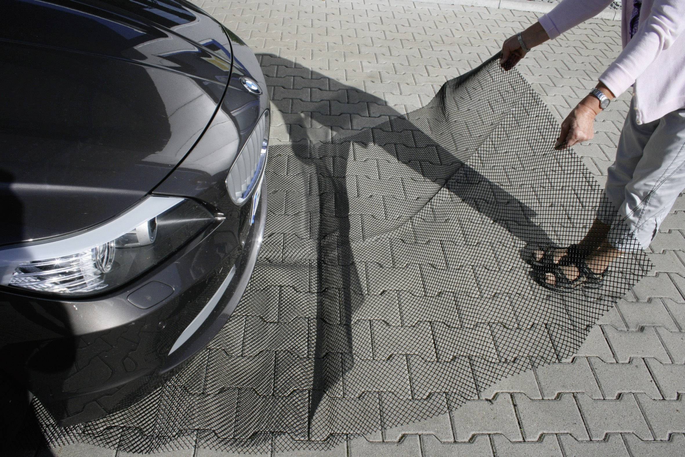Eine Person hält ein großes Gitter vor der Front eines Autos, während sie auf einer gepflasterten Fläche steht.