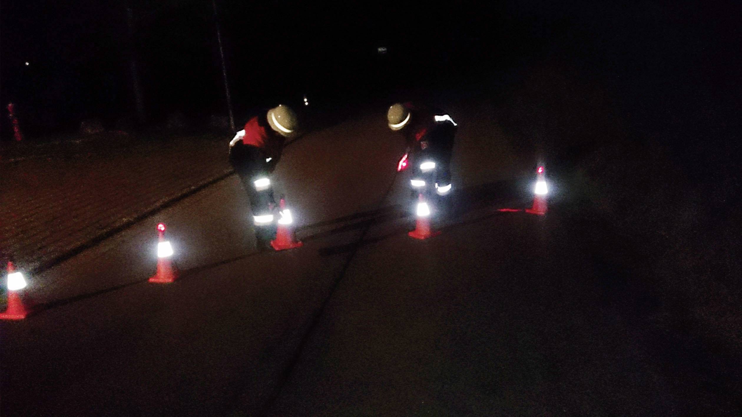 Zwei Personen in Schutzkleidung markieren nachts eine Straße mit reflektierenden Kegeln. Die Szene ist dunkel und schwer erkennbar.