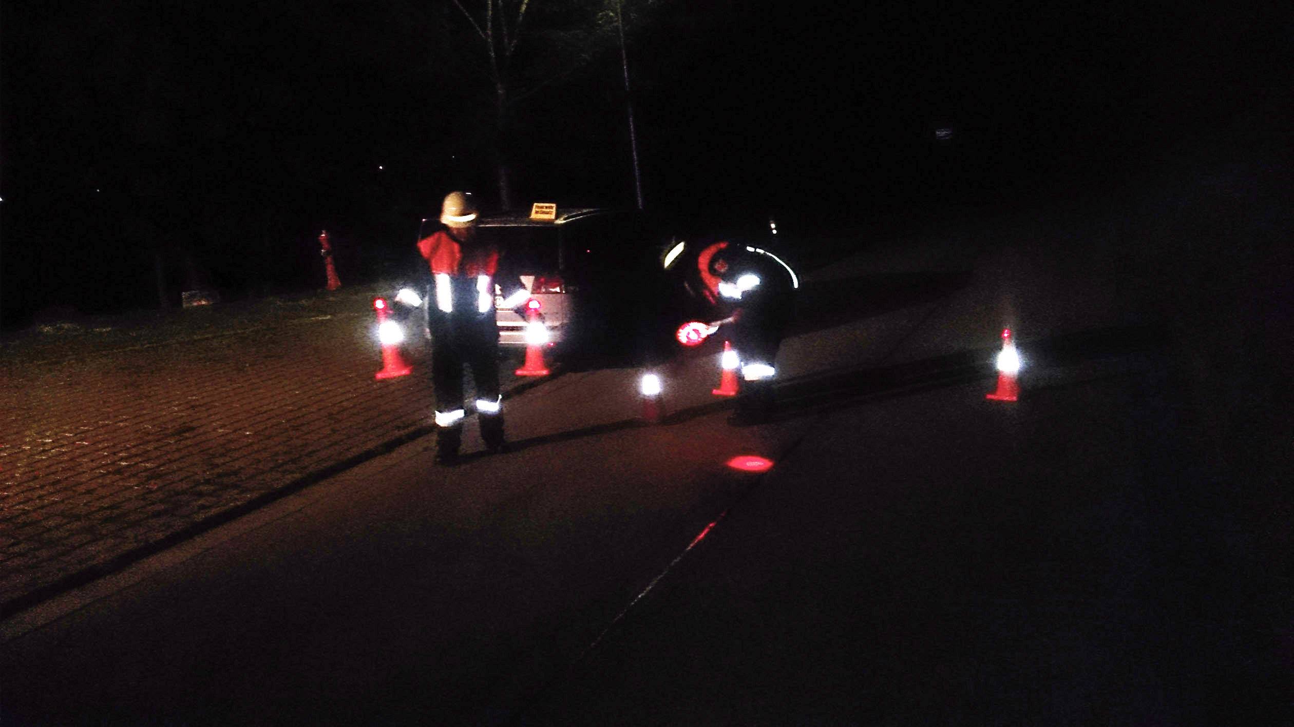 Zwei Personen in Sicherheitskleidung stehen nachts vor einem Auto. Sie sind von Verkehrsleitkegeln umgeben, möglicherweise bei einer Kontrolle.