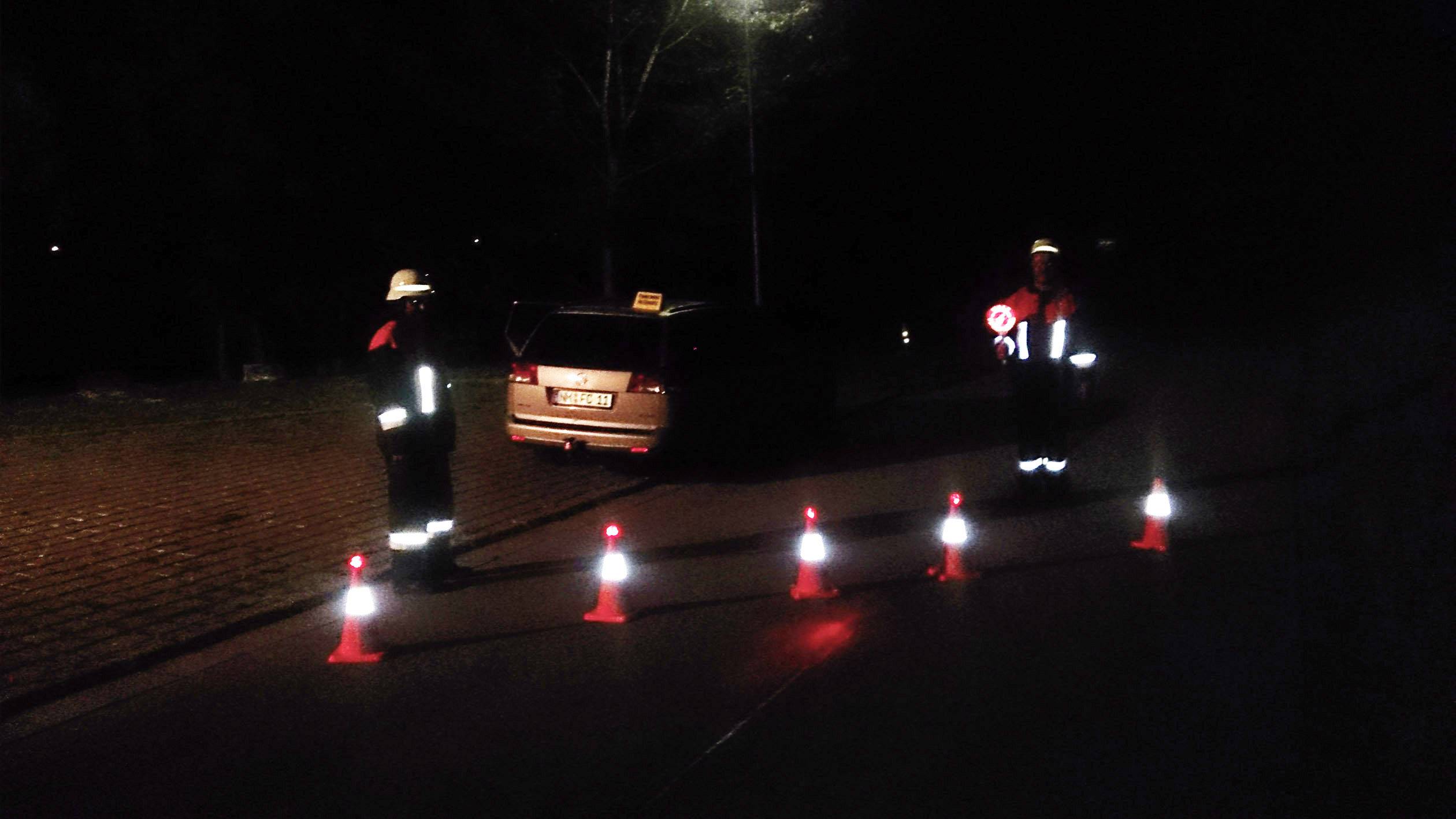 Zwei Personen in Warnwesten sichern bei Nacht eine Straße mit Absperrkegeln, ein Auto mit eingeschaltetem Warnblinker ist im Hintergrund.