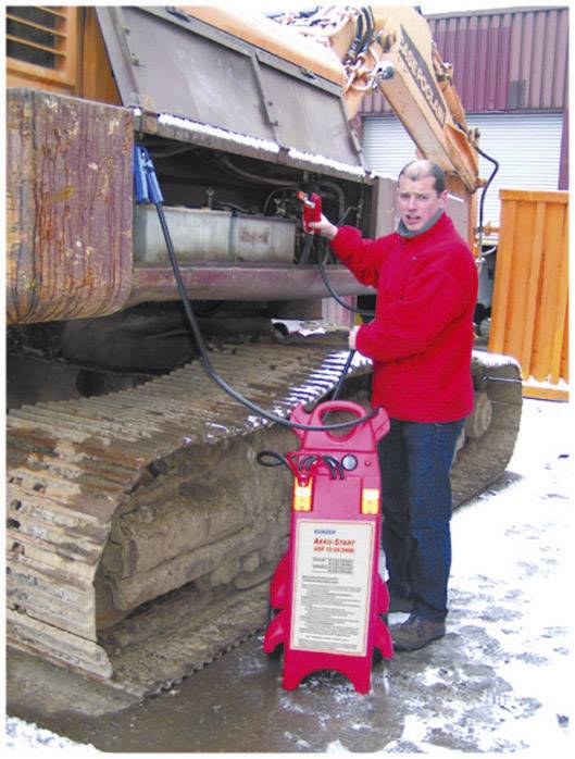Ein Mann in roter Jacke steht neben einem großen Baumaschine und füllt Flüssigkeit mit einer roten Apparatur nach. Schnee liegt auf dem Boden.