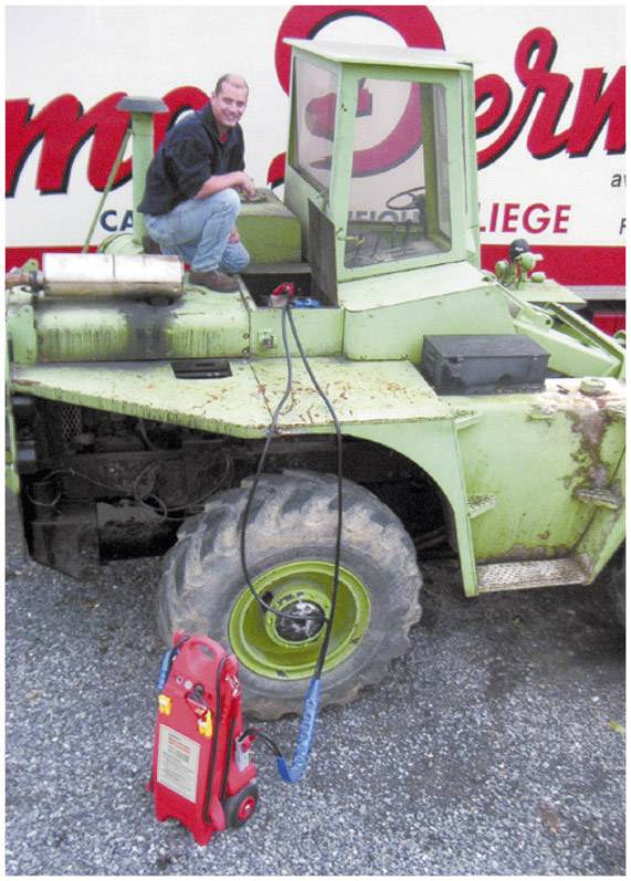 Ein Mann sitzt auf einem grünen Bagger neben einer roten Maschine. Im Hintergrund ist ein großes Fahrzeug mit Schriftzug sichtbar.