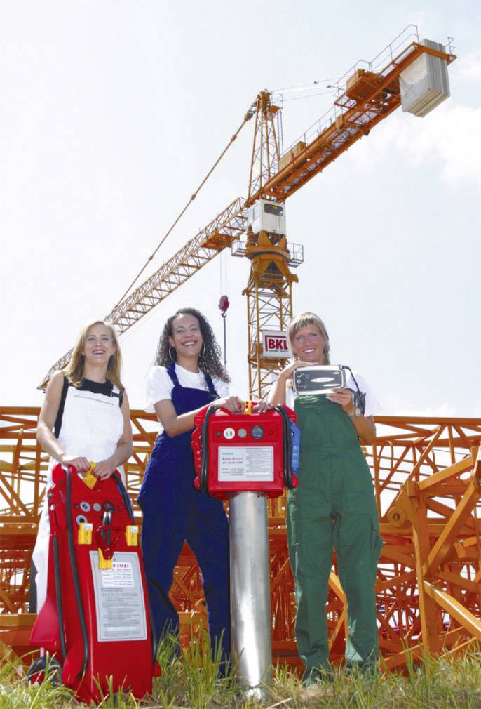 Drei Personen in Arbeitskleidung stehen vor einem großen Baukran auf einer Baustelle. Sie halten Sicherheitsausrüstung und Werkzeuge.