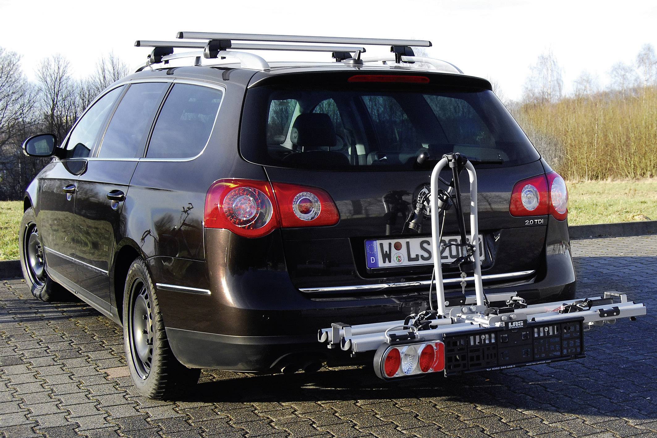 Ein dunkelbrauner Kombi mit einem Fahrradträger auf der Anhängerkupplung steht auf einem gepflasterten Parkplatz vor einem bewaldeten Hintergrund.