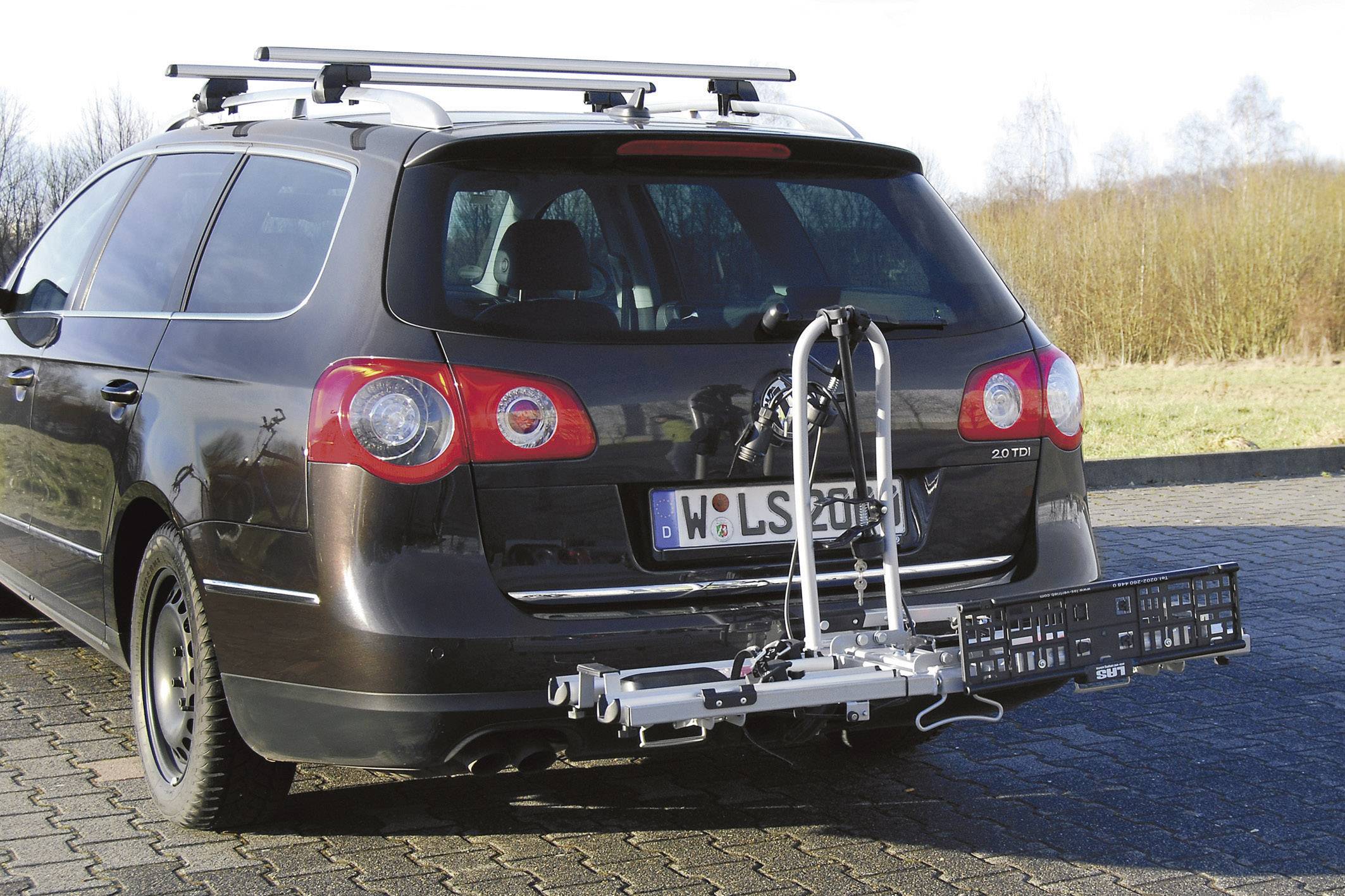 Auto mit abnehmbarem Fahrradträger am Heck, geparkt auf gepflastertem Platz. Träger ist leer, klarer Himmel und Bäume im Hintergrund.