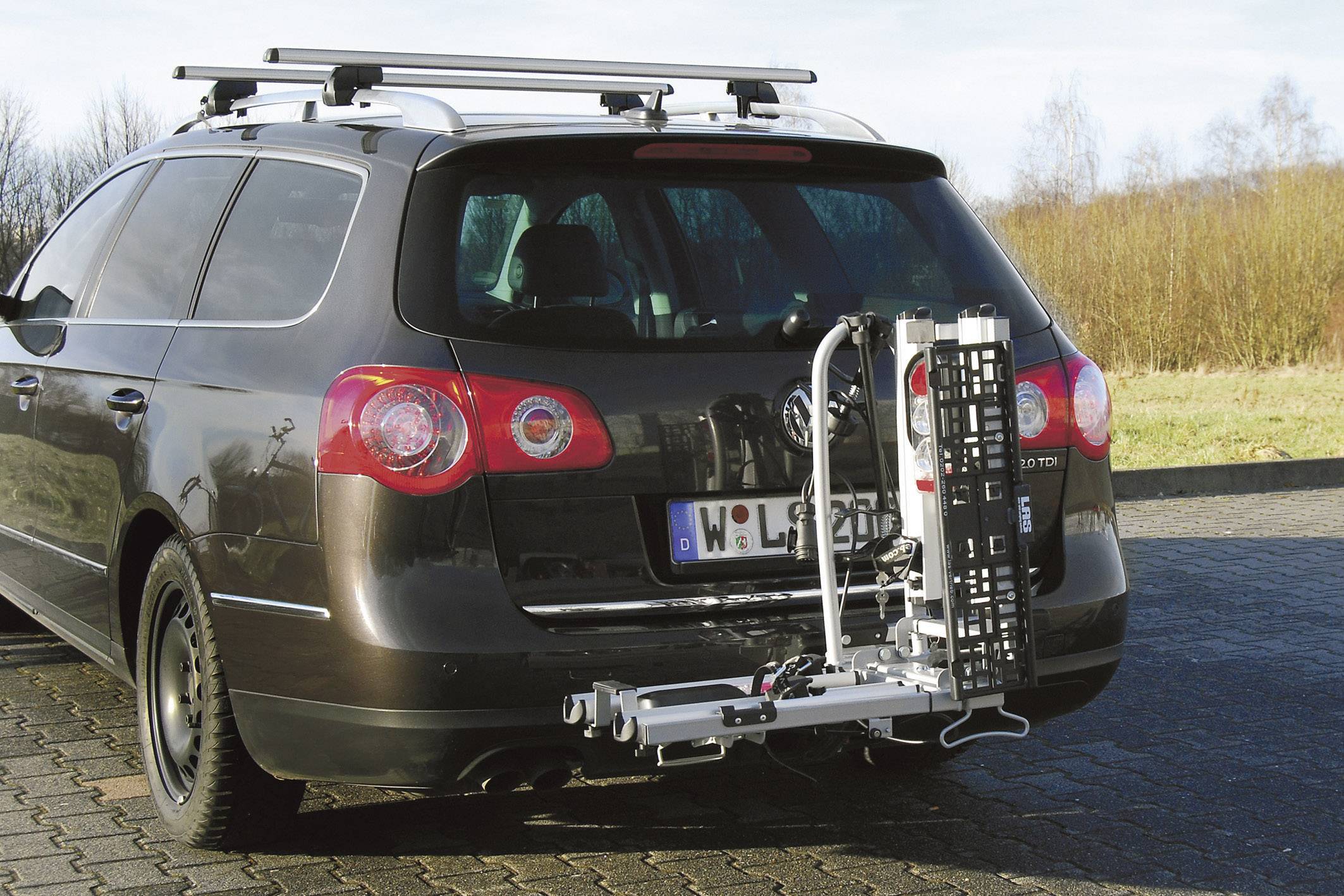 Ein dunkles Auto mit Dachgepäckträger hat ein metallisches Gestell am Heck befestigt. Es steht auf einem gepflasterten Platz.