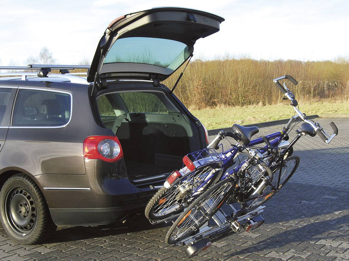 Ein offener Kombi-Kofferraum mit Fahrradträger und zwei Fahrrädern auf einem Parkplatz neben einer Wiese.