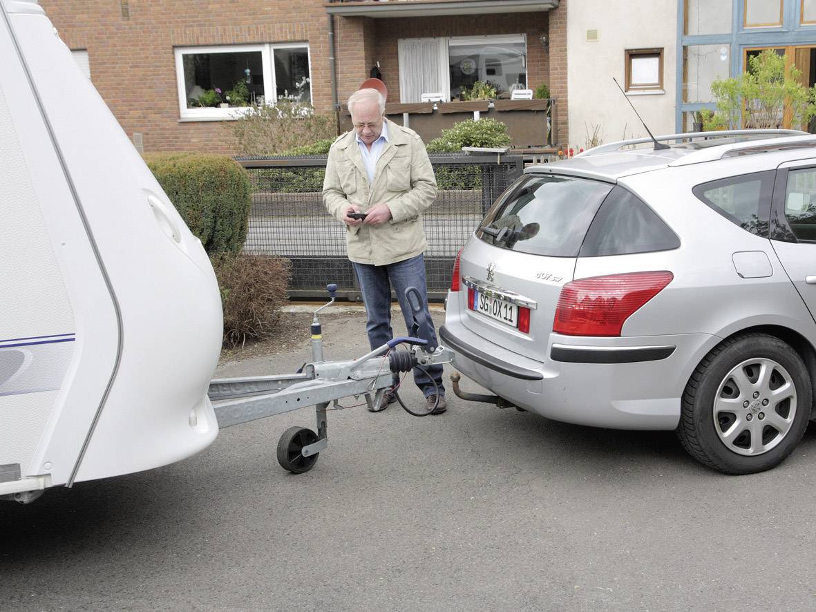 Ein Mann koppelt einen Wohnwagen an ein silbernes Auto an einer Wohnstraße. Er steht daneben, konzentriert auf die Verbindung.