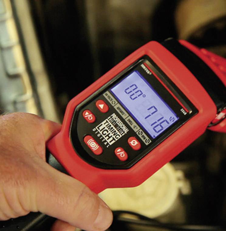 Eine Hand hält ein rotes digitales Diagnosegerät mit beleuchtetem Display, das '716' anzeigt, vor einem Hintergrund aus Autoteilen.