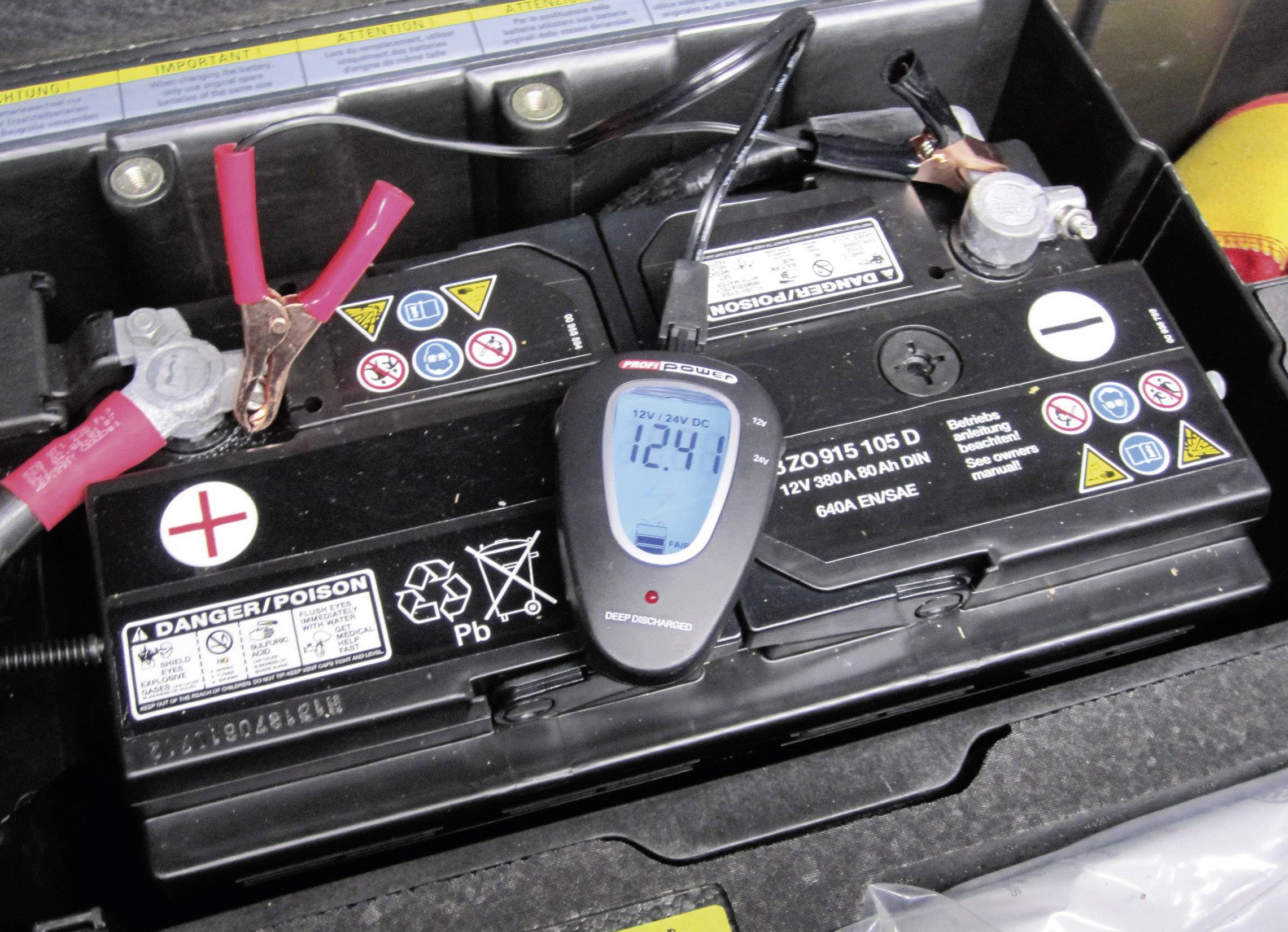 Autobatterie mit digitalem Voltmeter zeigt 12,4 Volt. Rote und schwarze Kabel sind an den Polen angeschlossen. Warnhinweise sichtbar.