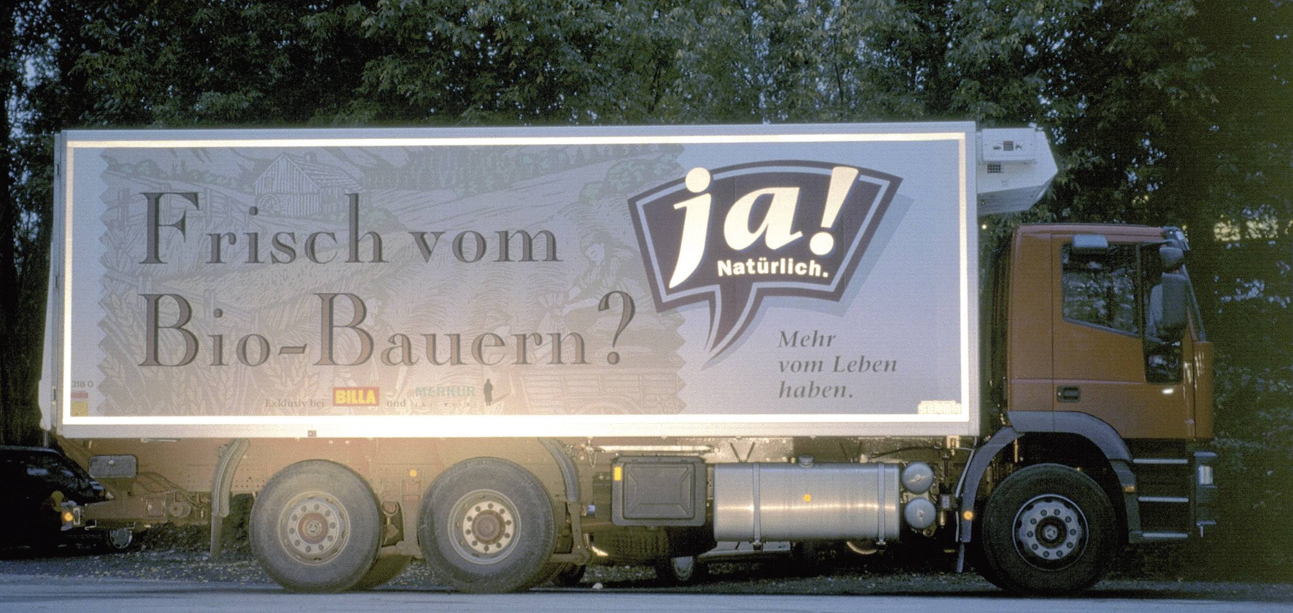 Ein Lastwagen mit der Aufschrift 'Frisch vom Bio-Bauern? Ja! Natürlich' steht auf einer Straße, umgeben von Bäumen.