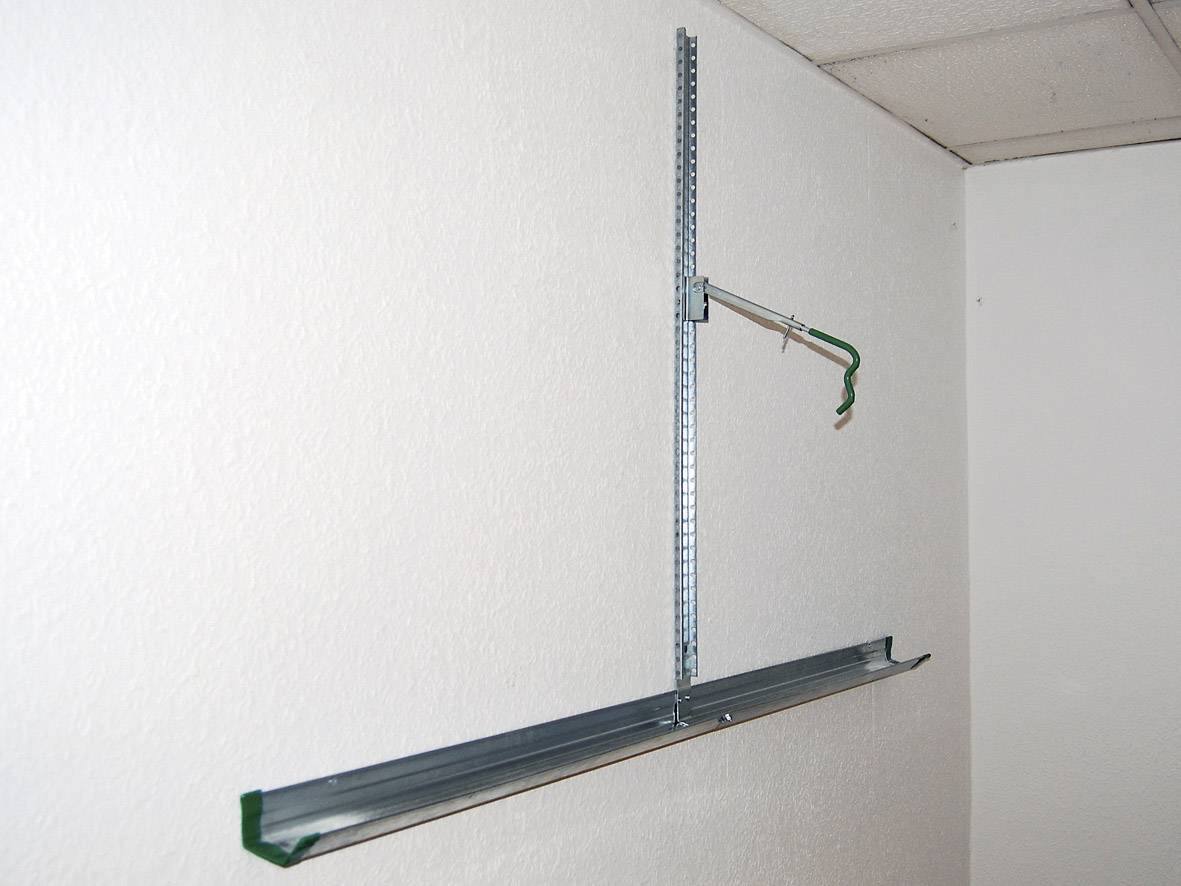 Ein Metallträger ist an einer Wand montiert. Ein weiteres Bauteil ist dazu befestigt und ein Haken ragt seitlich hervor.