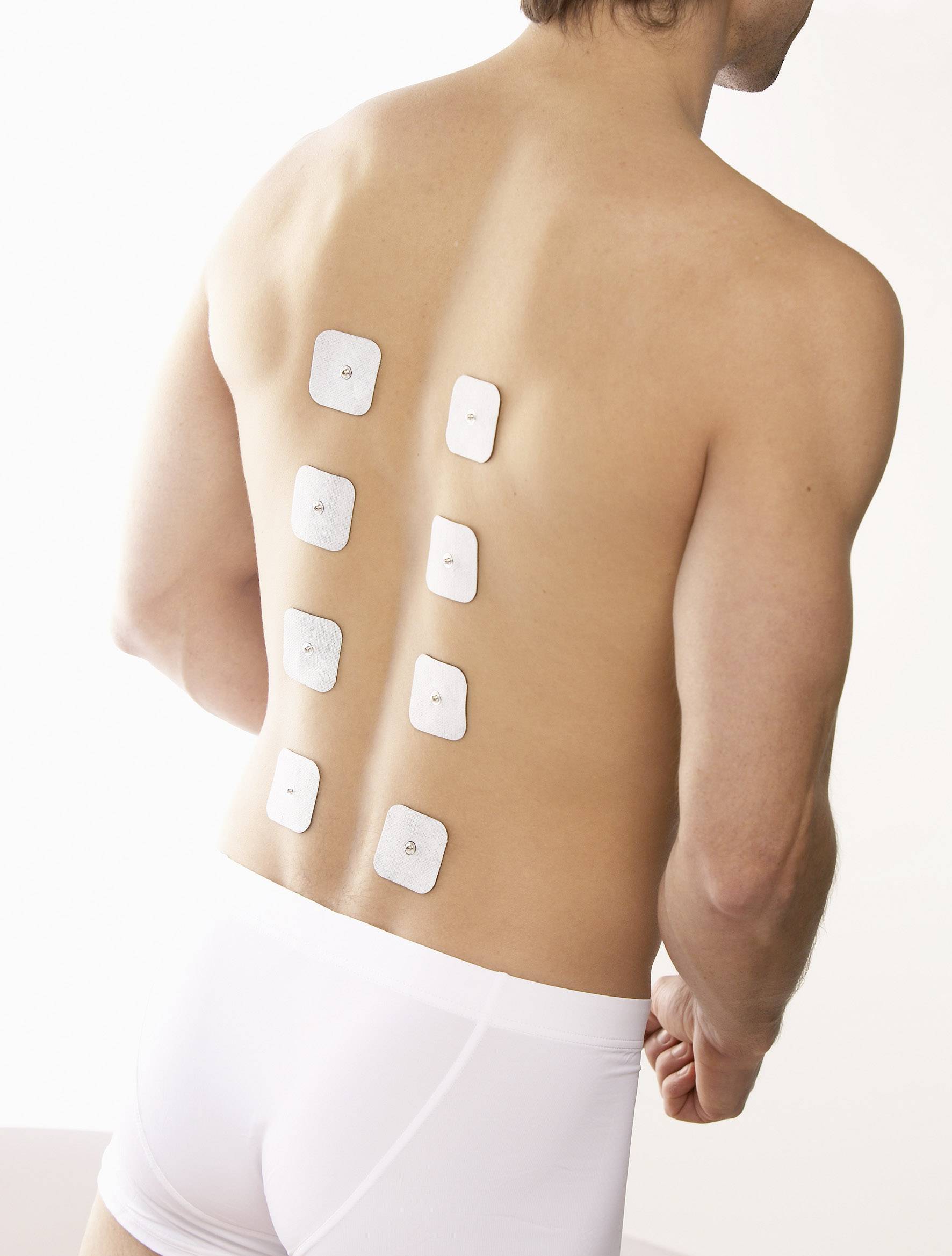 Eine Person von hinten mit Elektroden an Rücken, trägt weiße Unterwäsche, steht aufrecht, zeigt medizinische Behandlung oder Untersuchung.