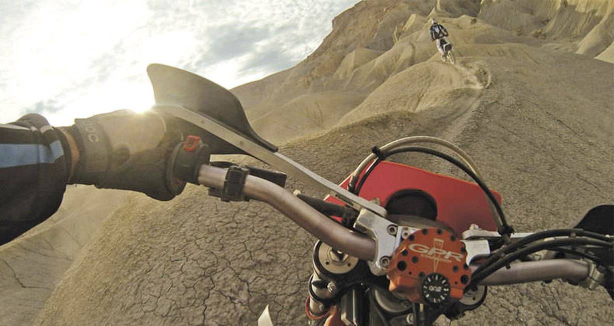 Motorradfahrer navigiert felsiges Gelände bei Sonnenuntergang, Berglandschaft im Hintergrund, Arm im Vordergrund sichtbar.