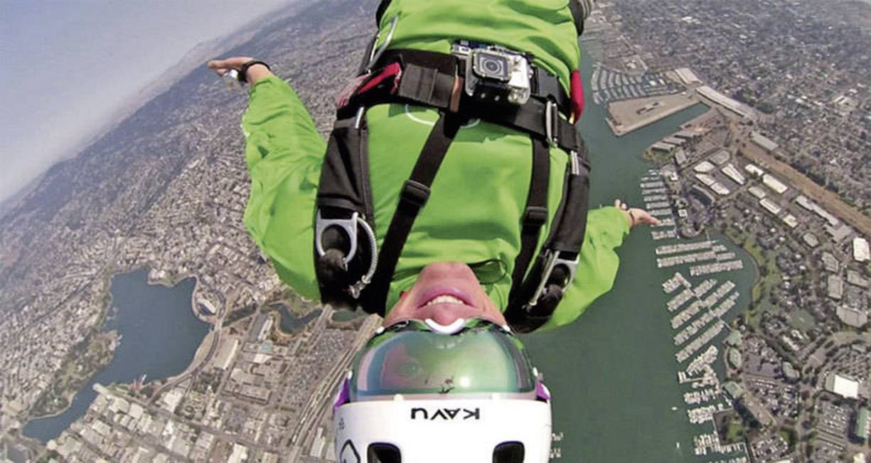 Eine Person im grünen Anzug springt mit einem Fallschirm von großem Höhen über einer Stadt mit Wasserwegen und Gebäuden.