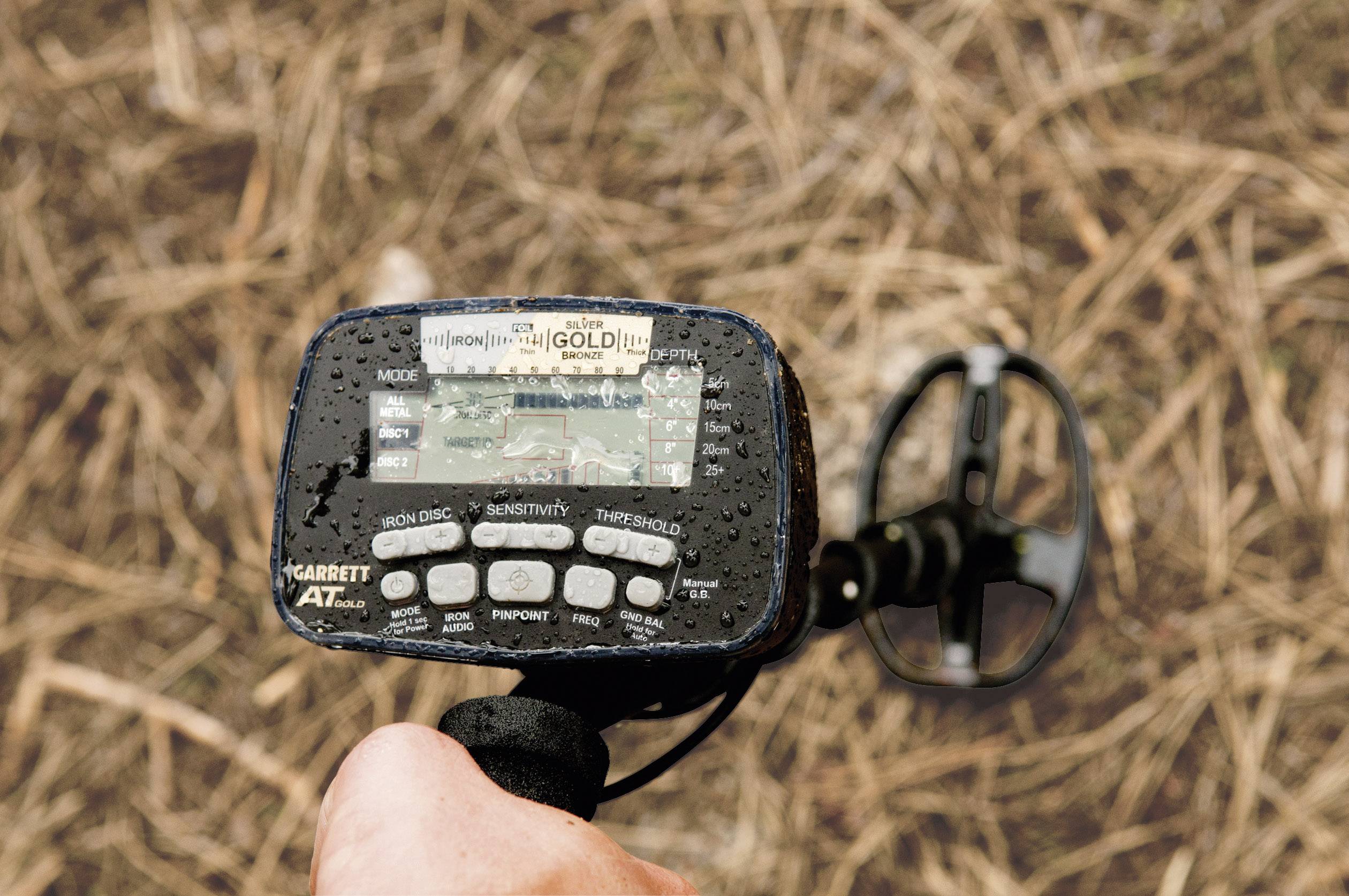 Eine Person hält einen Metalldetektor, der auf nassem Gras ausgerichtet ist. Der Bildschirm zeigt verschiedene Einstellungen.