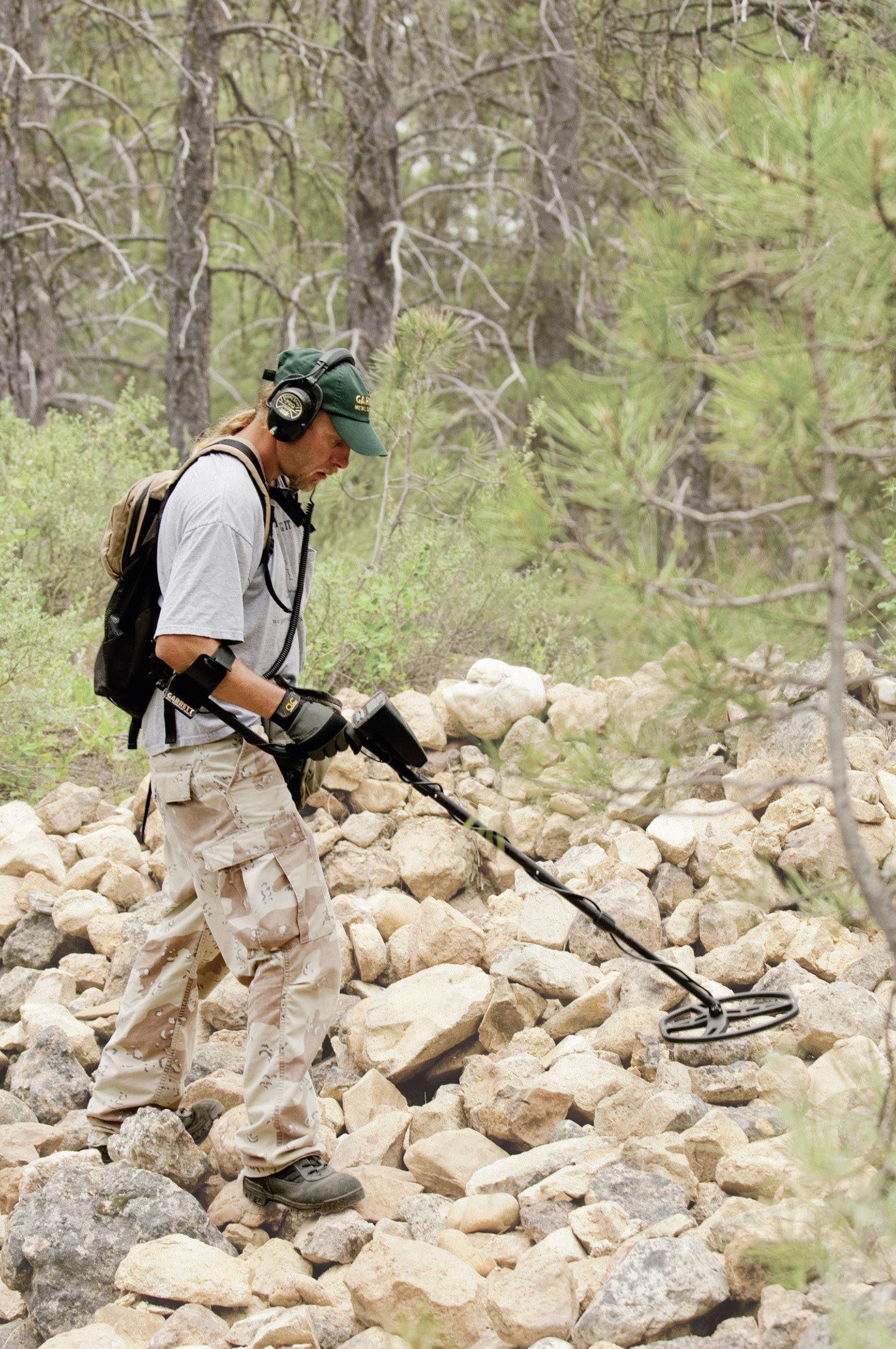 Ein Mann mit Kopfhörern benutzt einen Metalldetektor, um auf einem felsigen Pfad im Wald nach Metallen zu suchen.