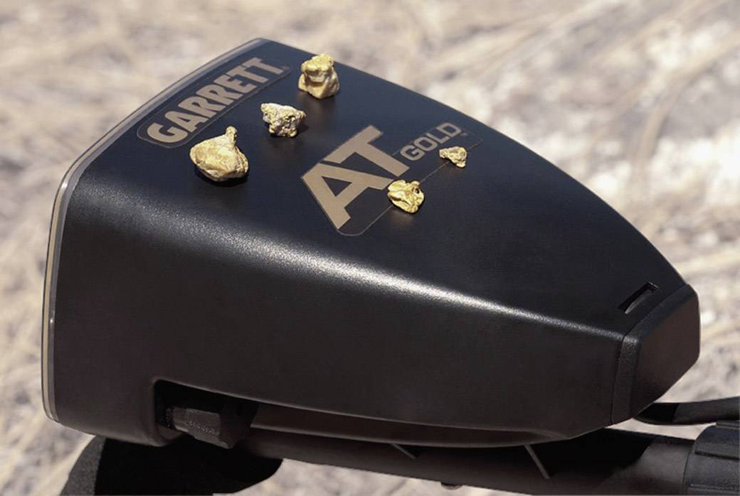 Metallsonde auf trockenem Boden, mit sechs kleinen Goldnuggets darauf. Der Markenname 'Garrett AT Gold' ist gut sichtbar.