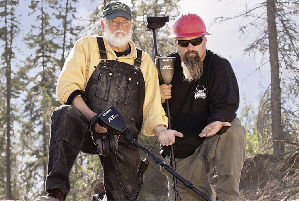 Zwei Personen mit Metalldetektoren in Outdoor-Kleidung knien auf Erde. Sie halten kleine Fundstücke in den Händen und blicken in die Kamera.