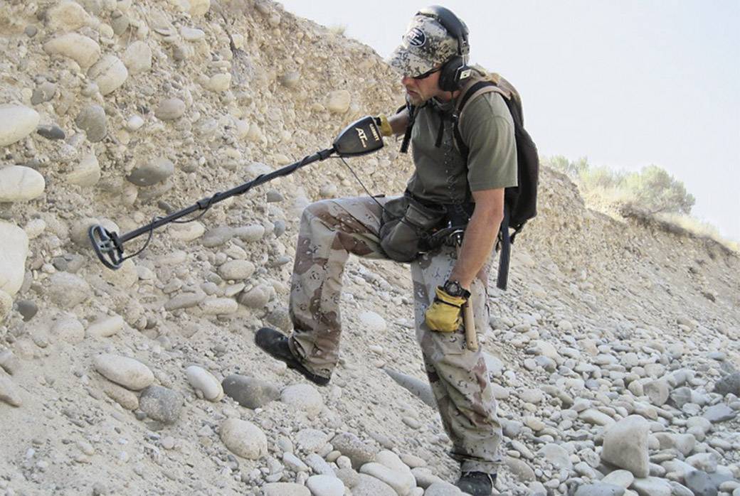 Eine Person mit Kopfhörern und einem Metalldetektor sucht in einer felsigen Wüstenlandschaft nach Metall.
