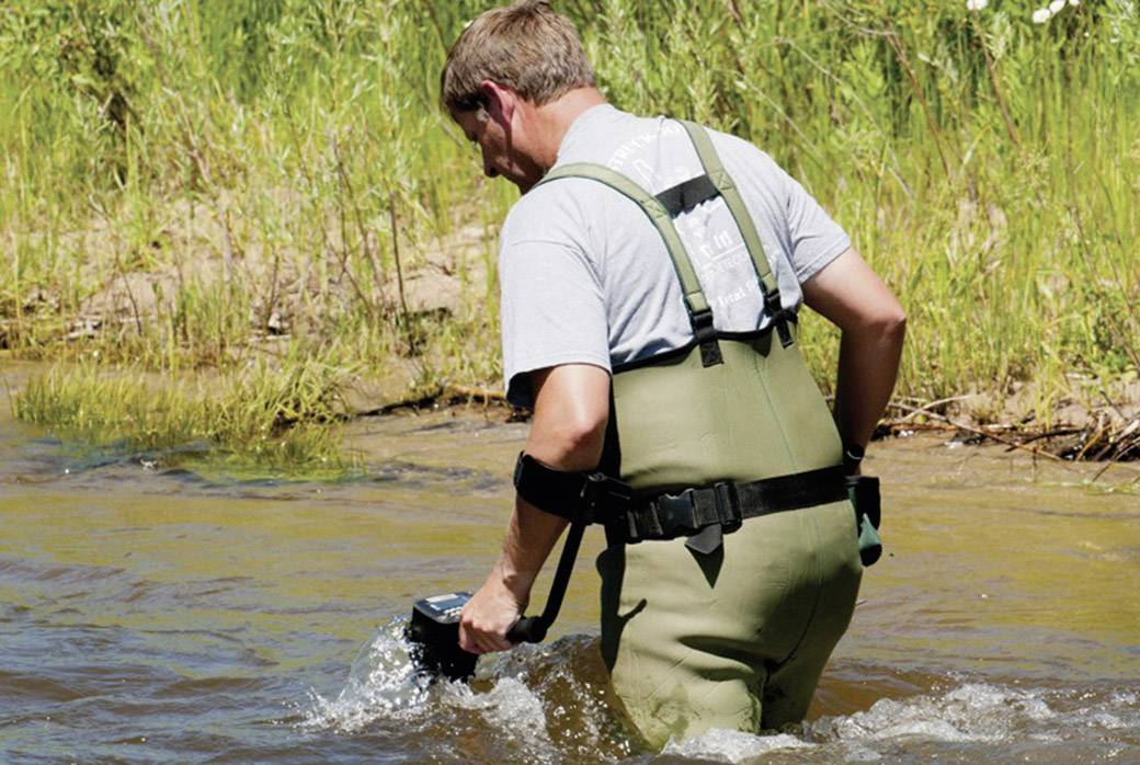Ein Mann watet in einem Fluss und misst die Wassertemperatur mit einem Gerät. Im Hintergrund sind Gras und Pflanzen sichtbar.