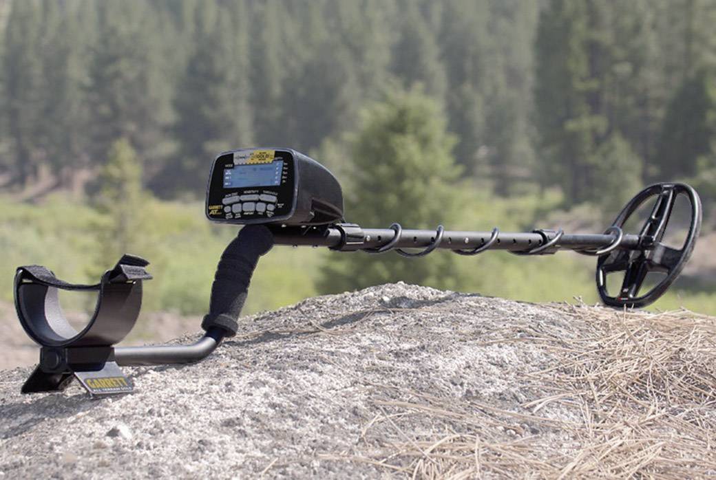 Metalldetektor ruht auf Felsen im Freien, umgeben von Bäumen. Gerät zeigt Display und Bedienelemente zur Metallsuche.