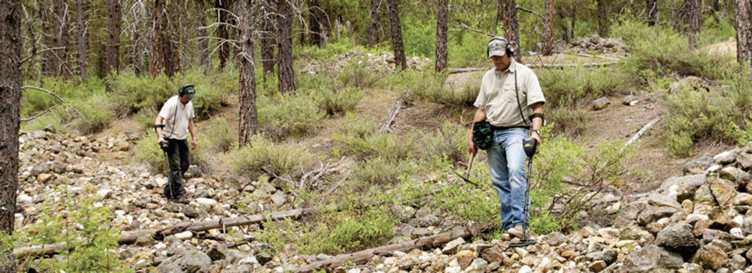 Zwei Personen gehen durch bewaldetes Gebiet mit steinigem Boden, umgeben von Bäumen und Sträuchern, bei Tageslicht.