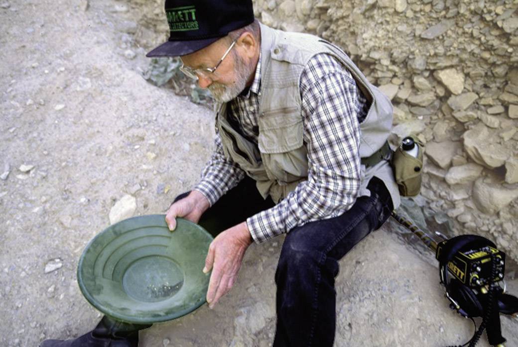 Ein älterer Mann sitzt in einer felsigen Umgebung und schwenkt eine grüne Pfanne, vermutlich zur Goldsuche, konzentriert und nachdenklich.
