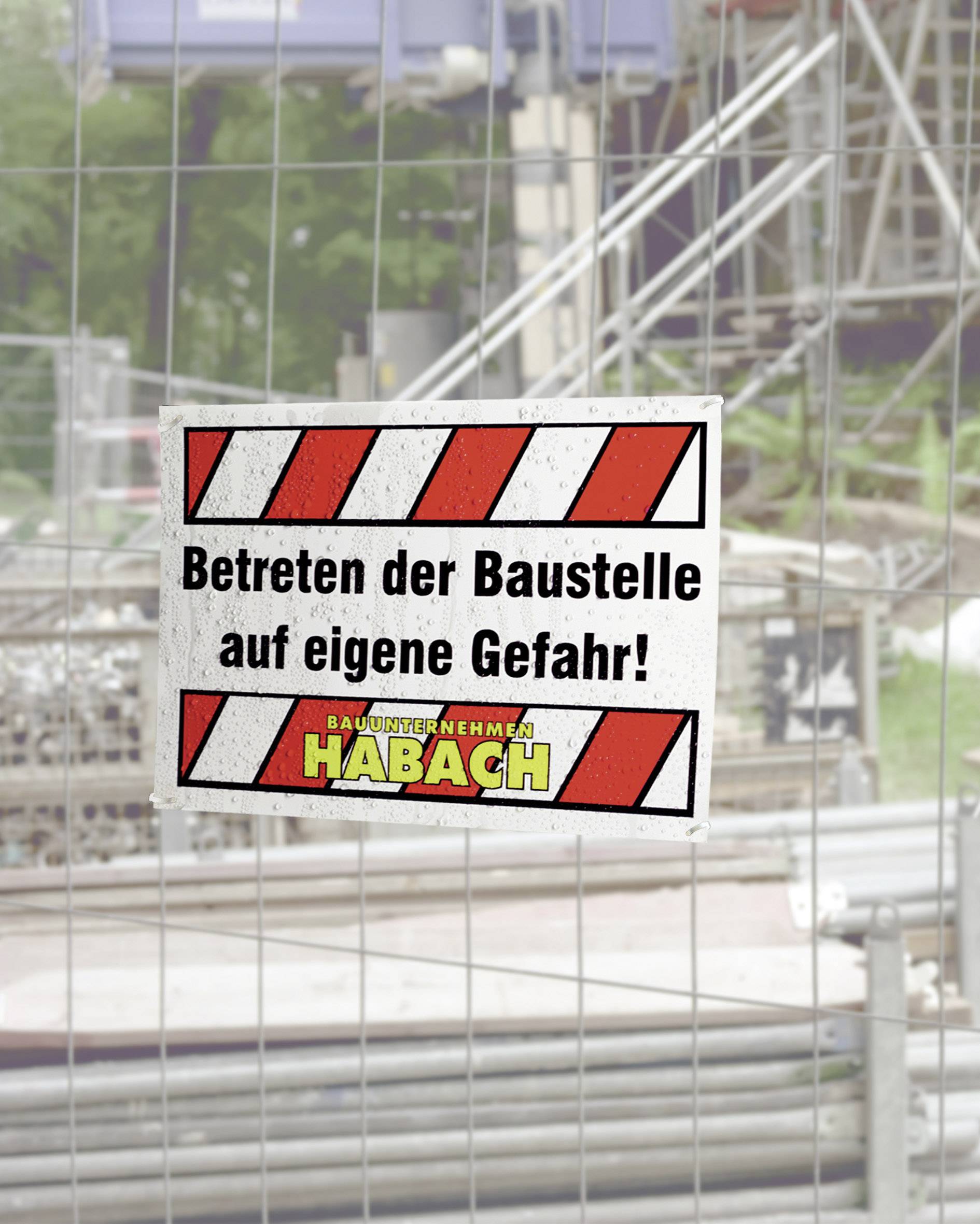 Ein Schild an einem Bauzaun warnt: 'Betreten der Baustelle auf eigene Gefahr! Bauunternehmen Habach.' Hintergrund zeigt Baustelle.