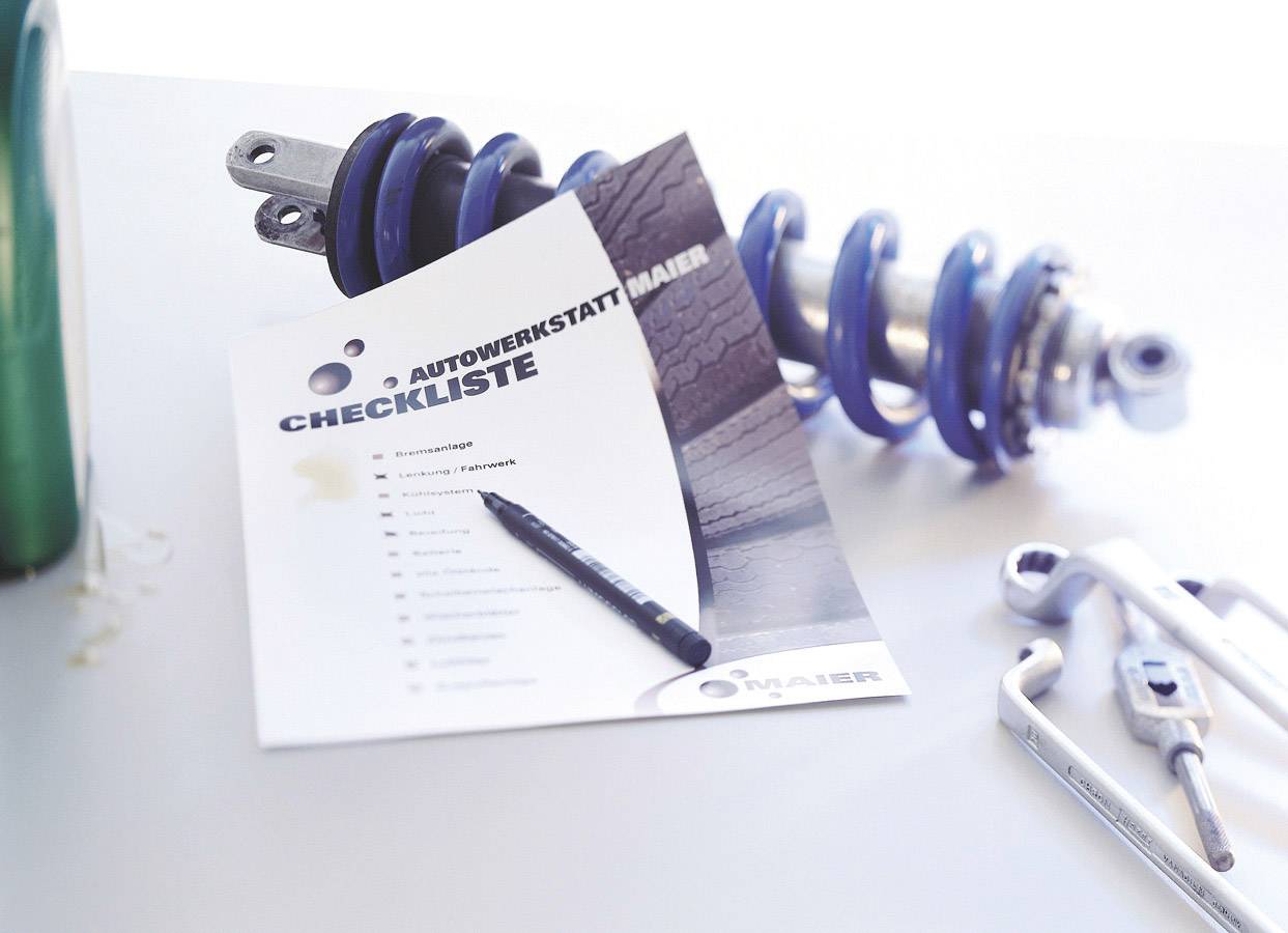 'Autowerkstatt-Checkliste' liegt neben Stoßdämpfern und Werkzeugen auf einem Tisch, zeigt Wartungsschritte in einer Werkstattumgebung.