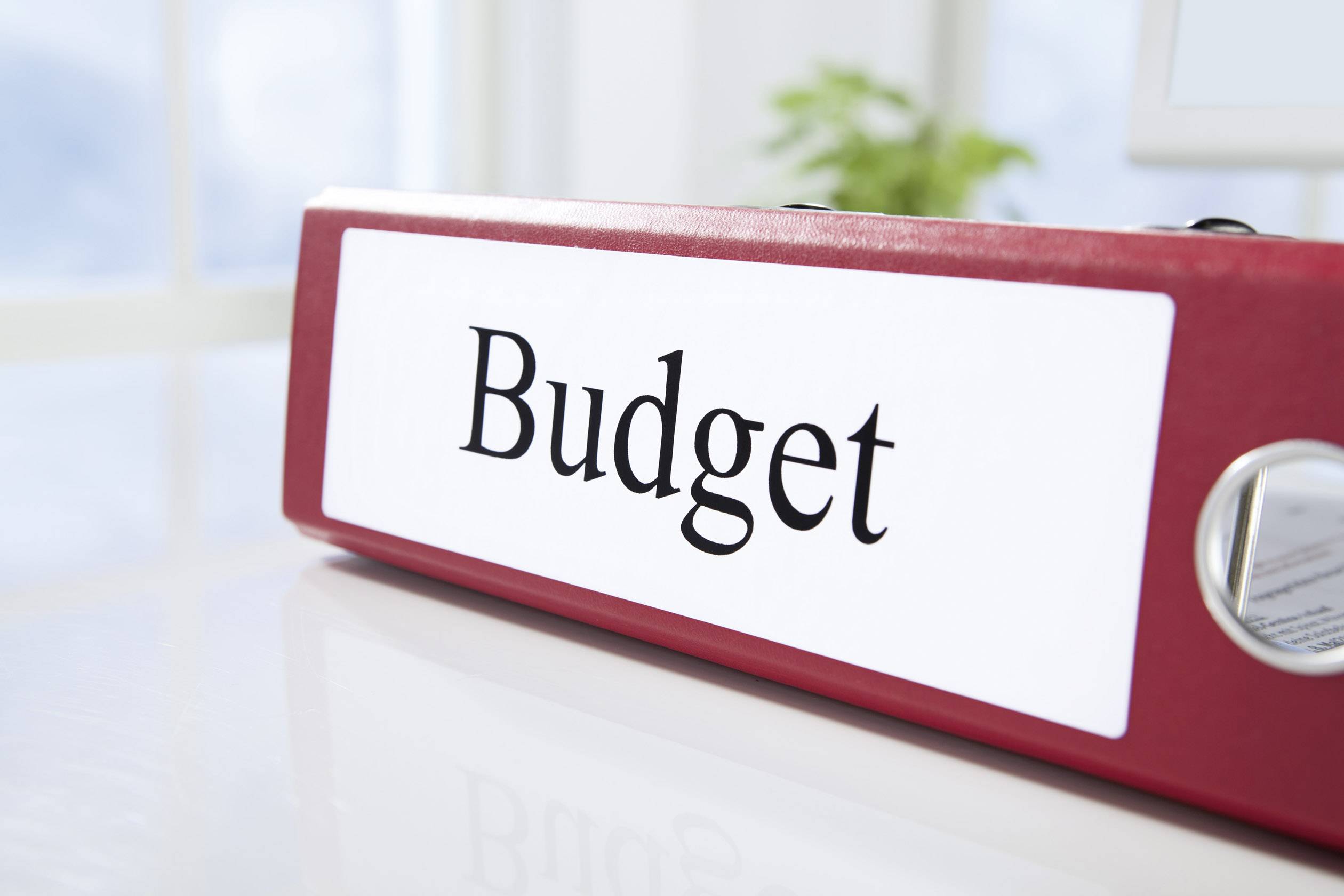 Ein roter Ordner mit der Aufschrift 'Budget' liegt auf einem Schreibtisch. Im Hintergrund sind unscharfe Fenster und Pflanzen zu sehen.
