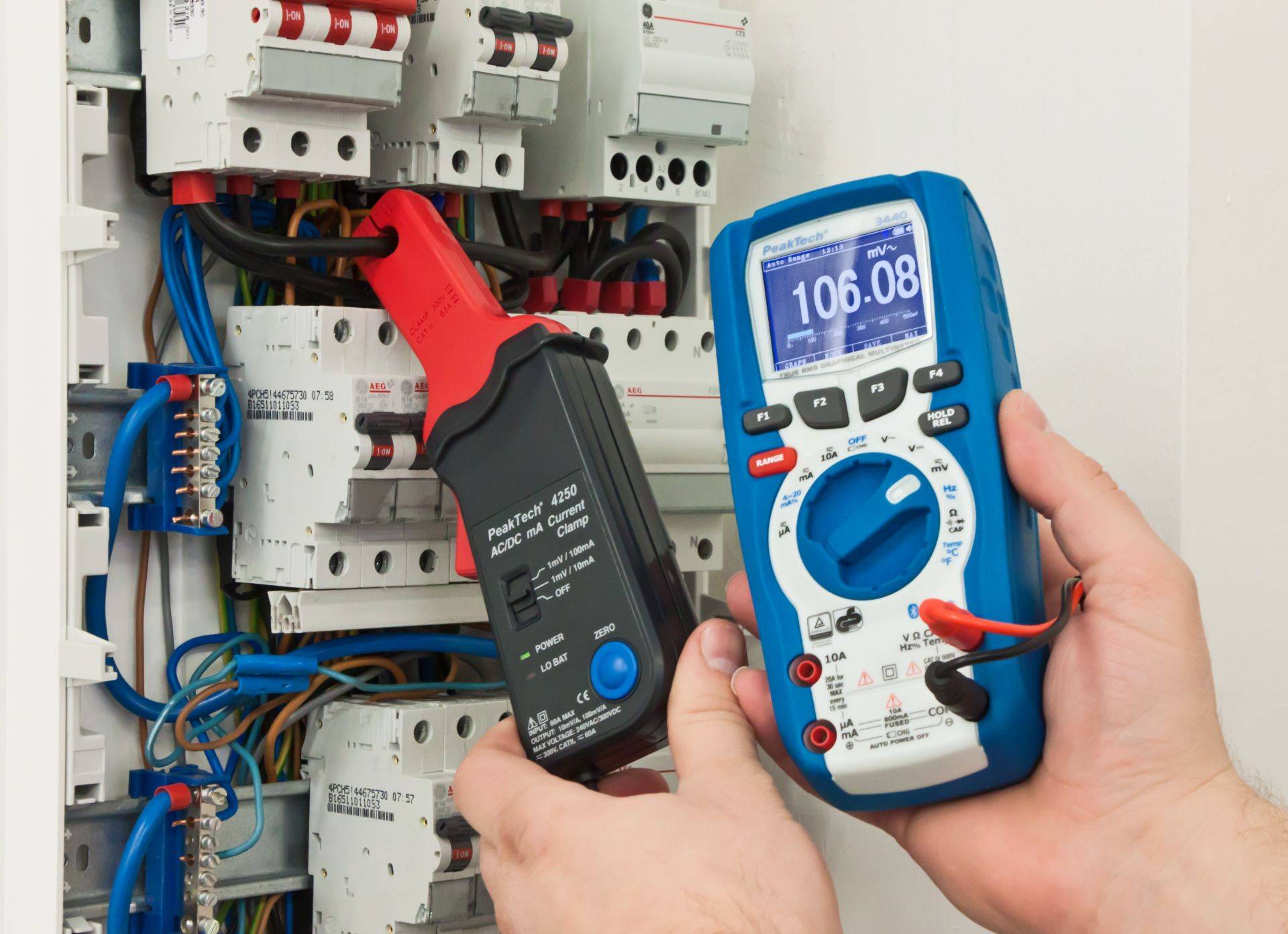 Zu sehen ist ein geöffneter Verteilerschrank mit mehreren Schaltern, die über Kabel miteinander verbunden sind. Eines der Kabel wird von einem schwarz-roten Stromzangenadapter umschlossen. Dieser ist mit einem blauen Digital-Multimeter verbunden. Beide Messgeräte werden von zwei Händen gehalten.