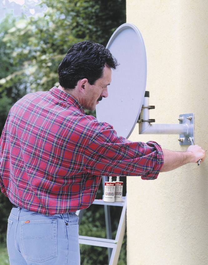 Ein Mann in kariertem Hemd montiert eine Satellitenschüssel an einer gelben Wand. Im Hintergrund sind Pflanzen zu sehen.
