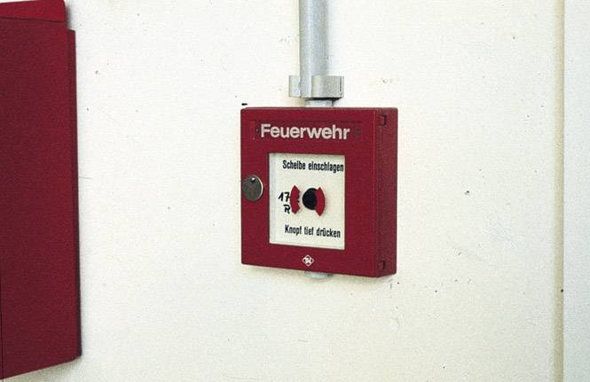 Roter Feuermelder an der Wand; Aufschrift 'Feuerwehr'. Anweisungen: 'Scheibe einschlagen, Knopf fest drücken'.