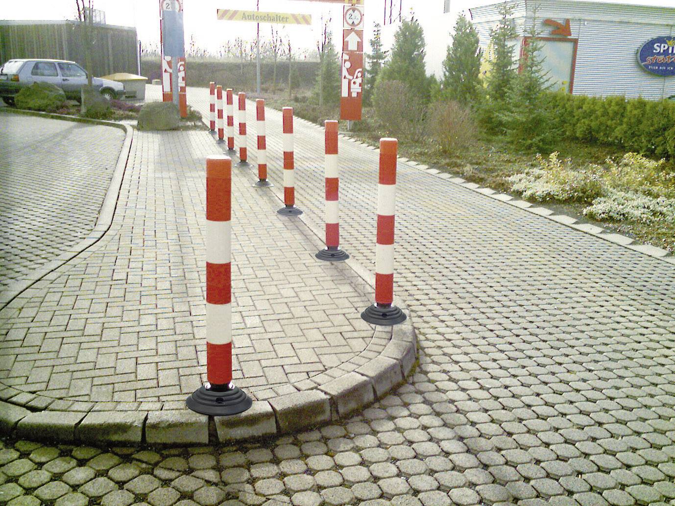 Mehrere rot-weiß gestreifte Poller auf einem gepflasterten Parkplatz markieren die Fahrbahn. Im Hintergrund Pflanzen und ein Gebäude.
