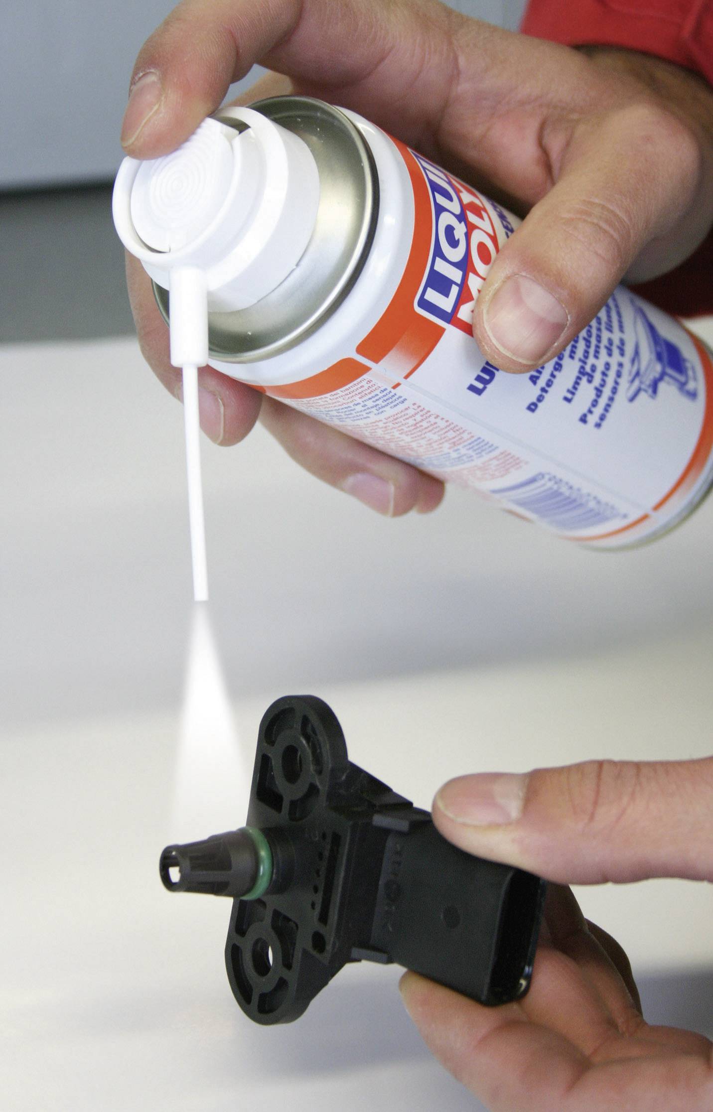 Eine Person sprüht Reiniger aus einer Dose auf einen kleinen schwarzen Sensor. Der Sensor wird in der Hand gehalten.